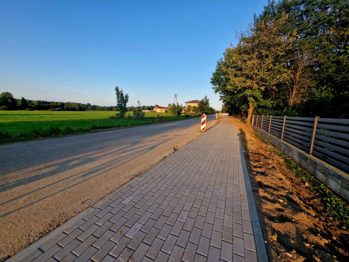 Droga z nowym chodnikiem z kostki brukowej, po prawej stronie ogrodzenie i drzewa, z lewej zielone pola. W tle niebo błękitne.