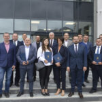 Grupa ludzi w formalnym ubraniu stoi na schodach przed dużym oknem. Trzymają niebieskie teczki. Tło to szklana fasada budynku.