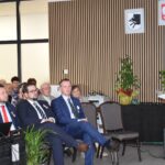 Grupa osób uczestniczy w spotkaniu w sali z drewnianymi panelami. Kobieta w bordowej sukience przemawia przy mikrofonie. W tle widać rośliny i symbole.