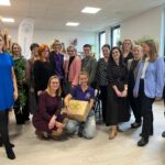 Grupa osób uśmiechniętych w bibliotece, stoją przed regałem z książkami. Osoba w niebieskiej sukience po lewej, reszta w eleganckich strojach. Jedna osoba klęczy z pudełkiem przewiązanym wstążką.