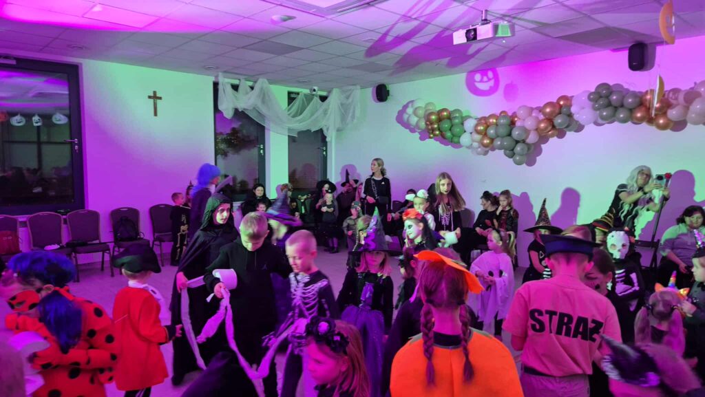 Na zdjęciu dzieci w kostiumach na imprezie halloweenowej. Jasne oświetlenie z fioletowymi i zielonymi akcentami. Tło zdobią balony i delikatne zasłony.