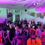 Na zdjęciu dzieci w kostiumach na imprezie halloweenowej. Jasne oświetlenie z fioletowymi i zielonymi akcentami. Tło zdobią balony i delikatne zasłony.