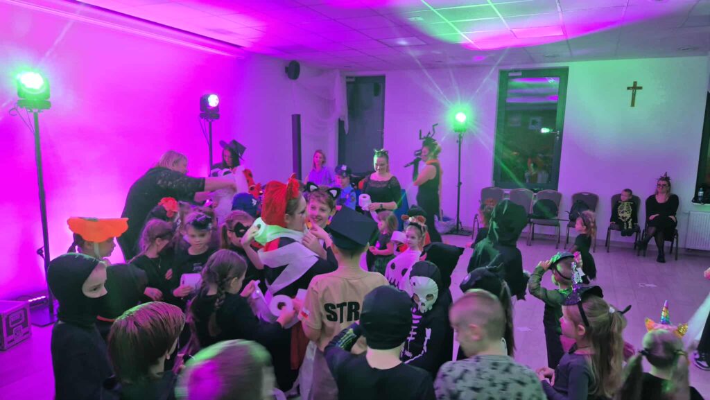 Grupa dzieci w kolorowych strojach bawi się na imprezie. Tło oświetlone jest fioletowymi światłami. Na ścianie wisi krzyż.