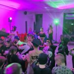 Grupa dzieci w kolorowych strojach bawi się na imprezie. Tło oświetlone jest fioletowymi światłami. Na ścianie wisi krzyż.