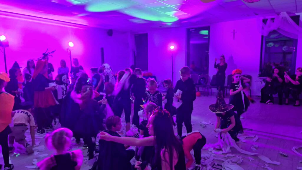 Widoczne jest halloweenowe przyjęcie dla dzieci w kolorowych kostiumach. Sala oświetlona na fioletowo z dekoracjami i papierem na podłodze. W tle są rodzice robiący zdjęcia.