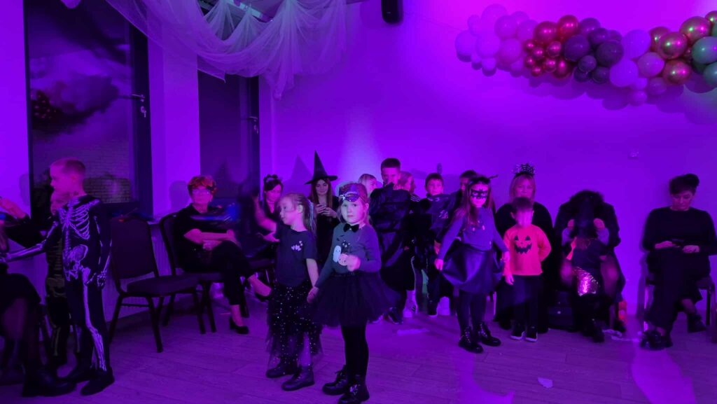 Dzieci w kostiumach stoją na fioletowo oświetlonej sali z balonami. Tło zdobią zasłony i ozdoby z białych i kolorowych balonów. Dorośli siedzą na krzesłach w tle.