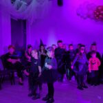 Dzieci w kostiumach stoją na fioletowo oświetlonej sali z balonami. Tło zdobią zasłony i ozdoby z białych i kolorowych balonów. Dorośli siedzą na krzesłach w tle.