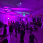 Sala udekorowana na Halloween, z dominacją fioletowego oświetlenia. Ludzie bawią się na parkiecie, w tle wiszą papierowe dynie. Kolorowe światła i halloweenowe motywy tworzą atmosferę zabawy.