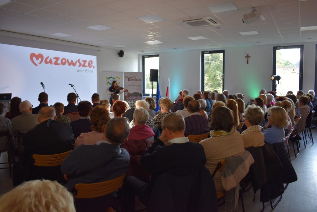 Sala z widownią siedzącą na krzesłach, w tle biały ekran z napisem Mazowsze. Osoby skupione na prezenterce obok mikrofonów.