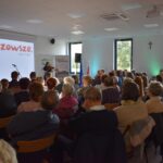 Sala z widownią siedzącą na krzesłach, w tle biały ekran z napisem Mazowsze. Osoby skupione na prezenterce obok mikrofonów.