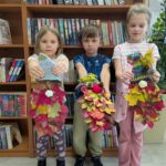 Troje dzieci trzyma przed sobą kolorowe, liściaste dekoracje. Stoją przed regałem pełnym książek. Tło jest jasne, z rośliną po prawej.
