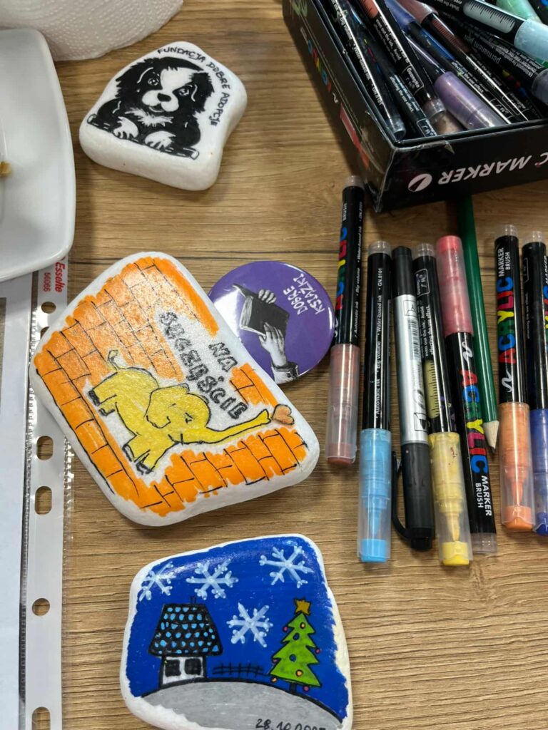 Na stole leżą pomalowane kamienie z motywami psa, słonia i zimowego krajobrazu. Kolory to czarny, pomarańczowy i niebieski. Obok znajduje się pudełko z pisakami.