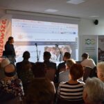 Osoba przemawia na tle prezentacji z logiem Uniwersytetu. Na scenie mikrofony, wokół siedzi publiczność. Z boku banery i światło z okna.