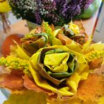 Bukiet z jesiennych liści w kształcie róż w odcieniach żółci i brązu. W tle fioletowy i biały wrzos. Drewniany stół pod spodem.