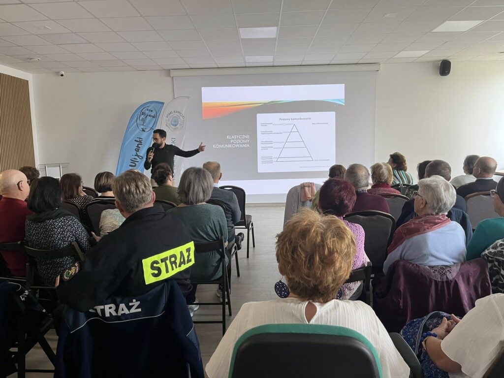 Mężczyzna prezentuje slajd o komunikacji na białym ekranie w sali. Uczestnicy siedzą na krzesłach, część w strojach z napisem STRAŻ. Jasne wnętrze z niebieskimi flagami w tle.