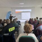 Mężczyzna prezentuje slajd o komunikacji na białym ekranie w sali. Uczestnicy siedzą na krzesłach, część w strojach z napisem STRAŻ. Jasne wnętrze z niebieskimi flagami w tle.