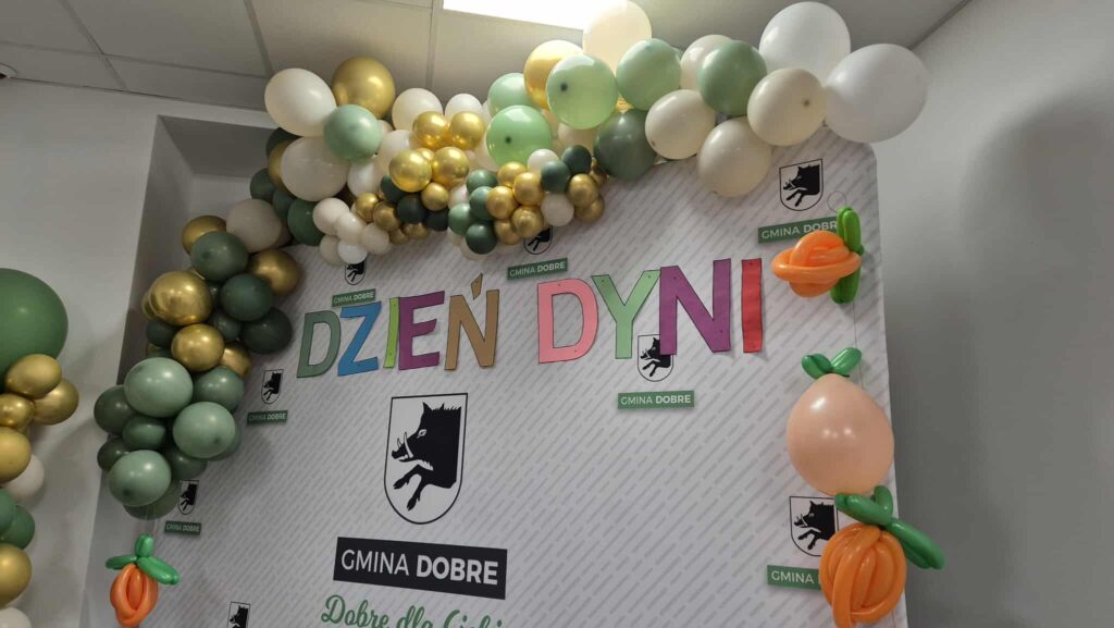 Na zdjęciu dekoracja z balonów w odcieniach zieleni, złota i bieli. Napis Dzień Dyni na białym tle. Ozdobne elementy przedstawiają dmuchane dynie.