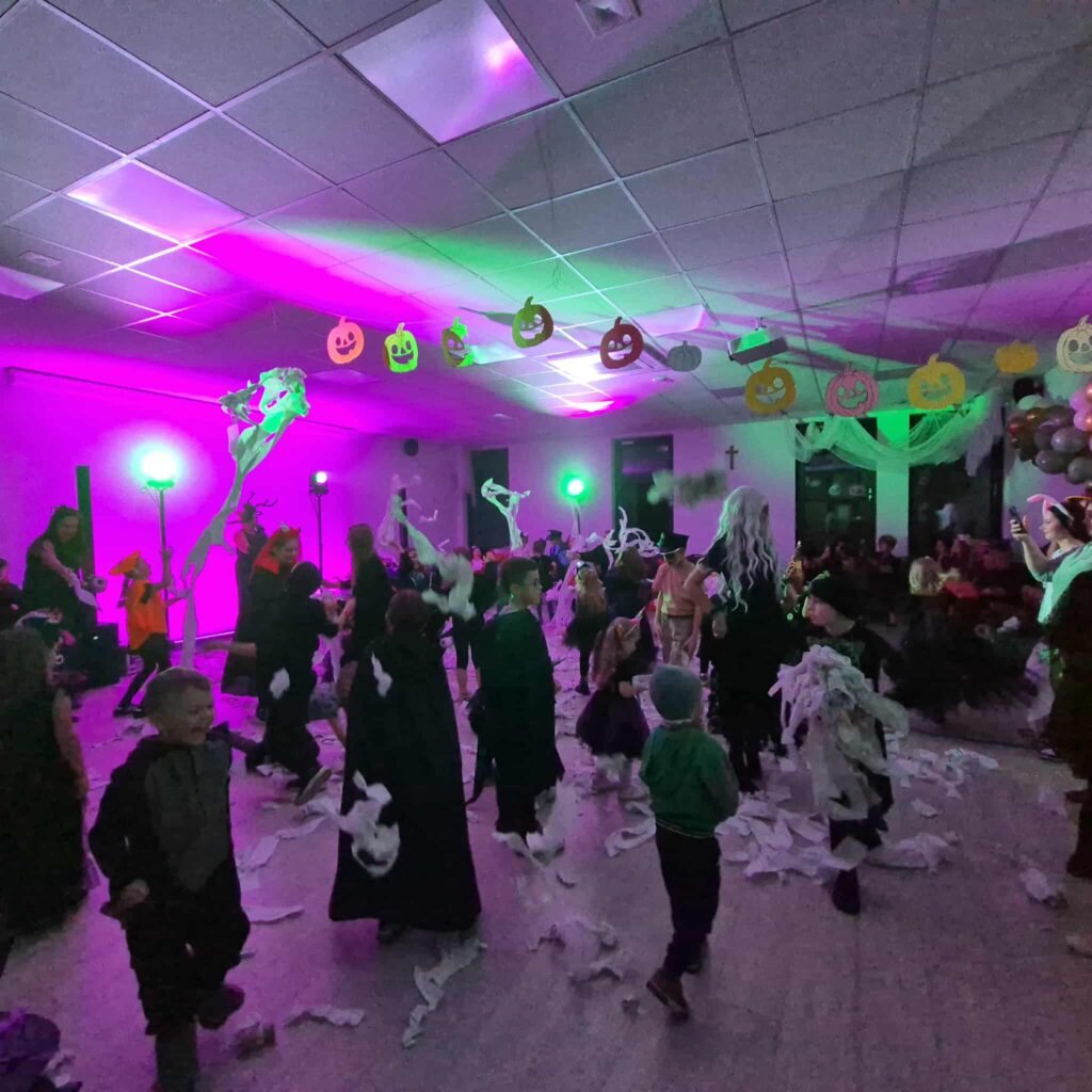 Pokój wypełniony dziećmi w kostiumach Halloween, otoczony zielonym i fioletowym światłem. Papier wiszący z sufitu. Dekoracje dyni na tle.