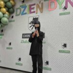 Na tle ściany z logo i napisem Dzień Dyni dziecko w czarnym stroju. Kolorowe balony w odcieniach zieleni. Tło szare z logo gminy.