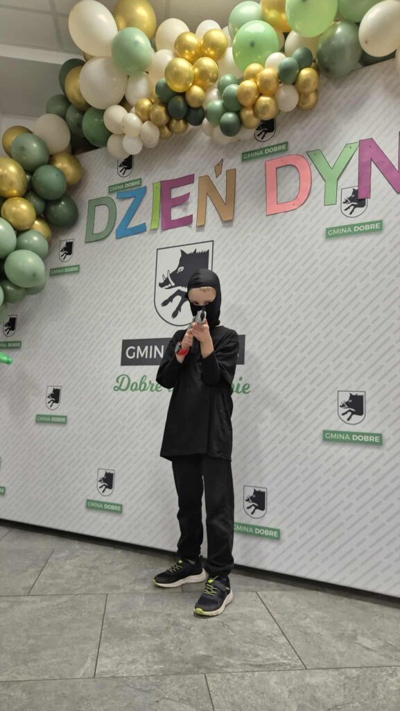 Na tle ściany z logo i napisem Dzień Dyni dziecko w czarnym stroju. Kolorowe balony w odcieniach zieleni. Tło szare z logo gminy.