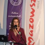 Na zdjęciu kobieta przemawia przy mikrofonie. W tle znajdują się fioletowy i biały baner z czerwonym napisem. Kobieta ma bordową sukienkę.