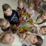 Dziewczynki trzymają kolorowe bukiety z liści. Stoją na jasnej podłodze. W tle widać półki pełne książek.