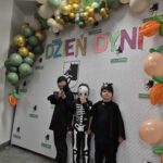 Trzech chłopców w kostiumach stoi przed ozdobną ścianką z balonami. Tło jest białe z kolorowym napisem „Dzień Dyni”. Balony są w odcieniach zieleni, złota i bieli.