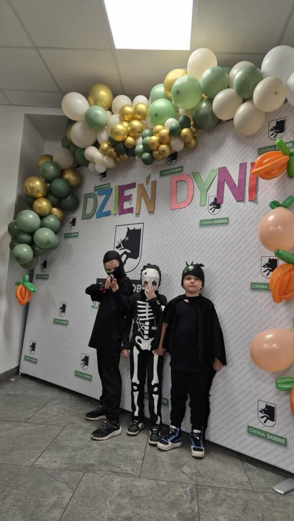 Trzech chłopców w kostiumach stoi przed ozdobną ścianką z balonami. Tło jest białe z kolorowym napisem „Dzień Dyni”. Balony są w odcieniach zieleni, złota i bieli.