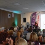 Na zdjęciu widoczna jest grupa ludzi siedzących na sali podczas prezentacji. Kobieta przy mikrofonie stoi na tle ekranu z logo. Ściany są jasne, z drewnianą dekoracją i plakatami.