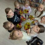 Grupa dzieci stoi na drewnianej podłodze, trzymając kolorowe bukiety. Dzieci są ubrane w jasne ubrania, a tło stanowi półka z książkami. Każde z nich patrzy w górę, do kamery.