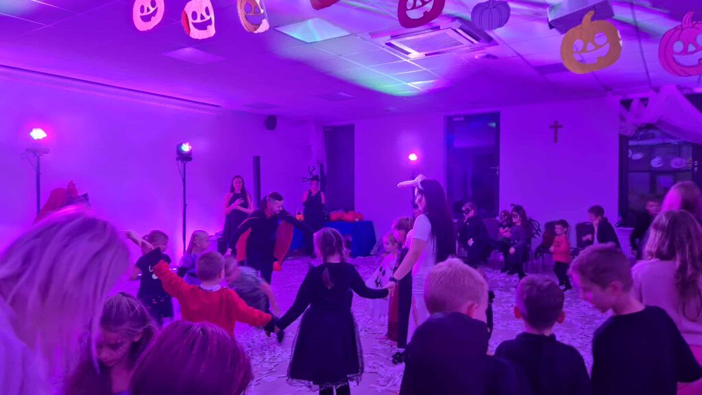 Na zdjęciu widać dzieci bawiące się na imprezie Halloween, w udekorowanej na fioletowo sali. Na suficie wiszą ozdobne dynie. W tle widać światła sceniczne i osoby dorosłe.