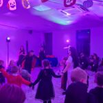 Na zdjęciu widać dzieci bawiące się na imprezie Halloween, w udekorowanej na fioletowo sali. Na suficie wiszą ozdobne dynie. W tle widać światła sceniczne i osoby dorosłe.