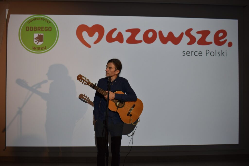 Osoba z gitarą stoi przed ekranem z logotypem Mazowsze i Uniwersytet Dobrego Wieku. Tło w kolorze białym. Widać cień postaci na ekranie.