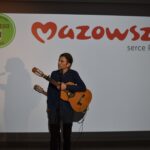 Osoba z gitarą stoi przed ekranem z logotypem Mazowsze i Uniwersytet Dobrego Wieku. Tło w kolorze białym. Widać cień postaci na ekranie.
