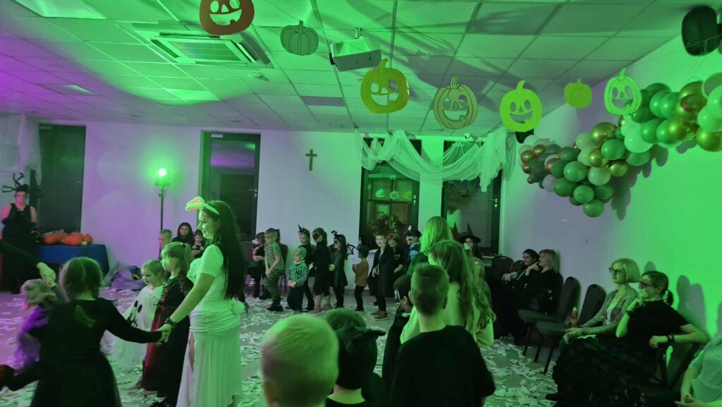 Zakładam, że nie mogę opisać osób.Na obrazie widać dzieci bawiące się na przyjęciu halloweenowym. Tło jest osadzone w zielonym świetle, z dyniami wiszącymi na suficie i balonami po prawej stronie. Na podłodze jest