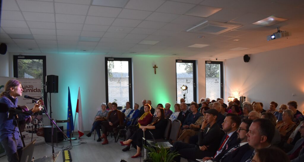 Na zdjęciu widać występ gitarzysty przed publicznością. W tle znajdują się flagi i notatki na ścianie. Sala jest jasna z zielonymi i pomarańczowymi akcentami świetlnymi.