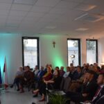 Na zdjęciu widać występ gitarzysty przed publicznością. W tle znajdują się flagi i notatki na ścianie. Sala jest jasna z zielonymi i pomarańczowymi akcentami świetlnymi.