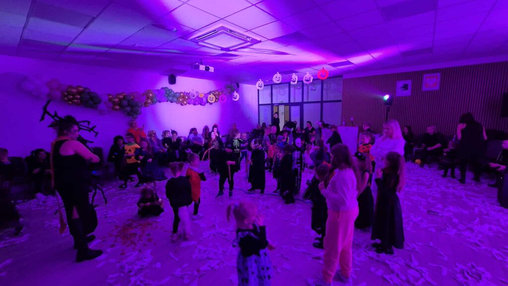 Na zdjęciu widać dzieci przebrane w kostiumy na balu przebierańców. Tło jest fioletowe z girlandą z balonów i wiszącymi dekoracjami. Podłoga jest pokryta białymi skrawkami papieru.