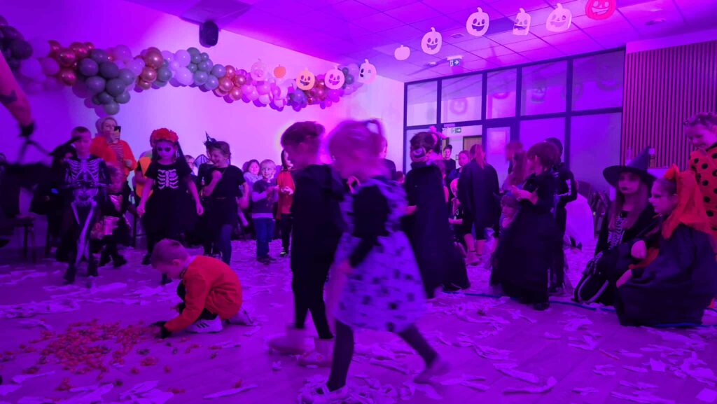 Dzieci w strojach Halloween bawią się w fioletowym oświetleniu. W tle balony i papierowe dynie na suficie. Na podłodze leżą rozrzucone papierki.