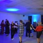 Grupa osób tańczących w jasno oświetlonej sali z niebieskim światłem. Tło z bannerem Mazowsze. Ubrania w odcieniach czerwieni i czerni.