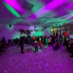 Pokój pełen dzieci w kostiumach z okazji Halloween. Fioletowe i zielone światła tworzą atmosferyczne efekty. Dekoracje z balonów i papierowe ozdoby pokrywają podłogę.