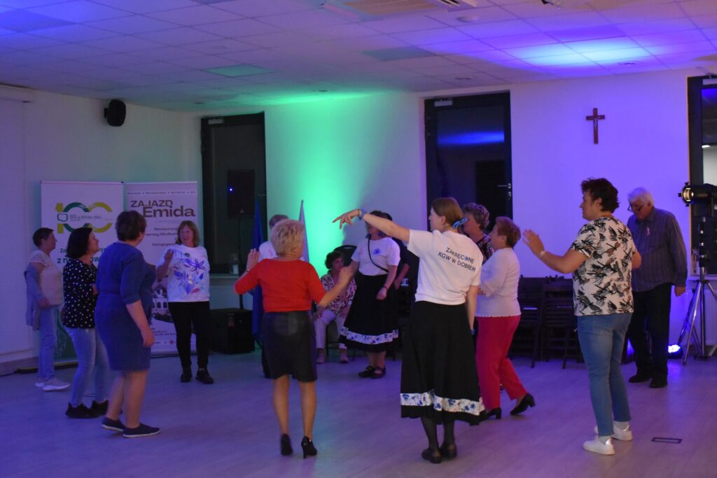 Grupa osób tańczy w jasnym pomieszczeniu z niebieskim oświetleniem. Kobieta w centrum nosi biały T-shirt z napisem. W tle banery i krzyż na ścianie.