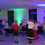 Grupa osób tańczy w jasnym pomieszczeniu z niebieskim oświetleniem. Kobieta w centrum nosi biały T-shirt z napisem. W tle banery i krzyż na ścianie.