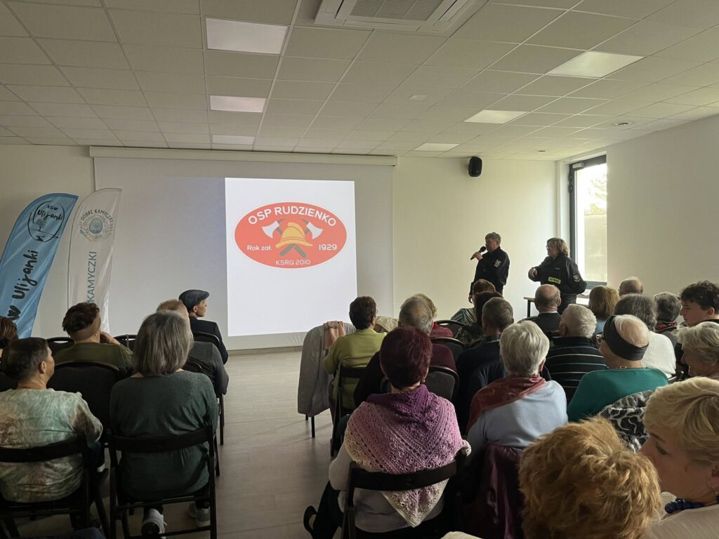 Sala zapełniona osobami siedzącymi, w tle ekran z logo OSP Rudzinko. Dwie osoby prowadzą prezentację przy mikrofonie. Zdjęcie wykonane w jasnym pomieszczeniu.