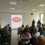 Sala zapełniona osobami siedzącymi, w tle ekran z logo OSP Rudzinko. Dwie osoby prowadzą prezentację przy mikrofonie. Zdjęcie wykonane w jasnym pomieszczeniu.