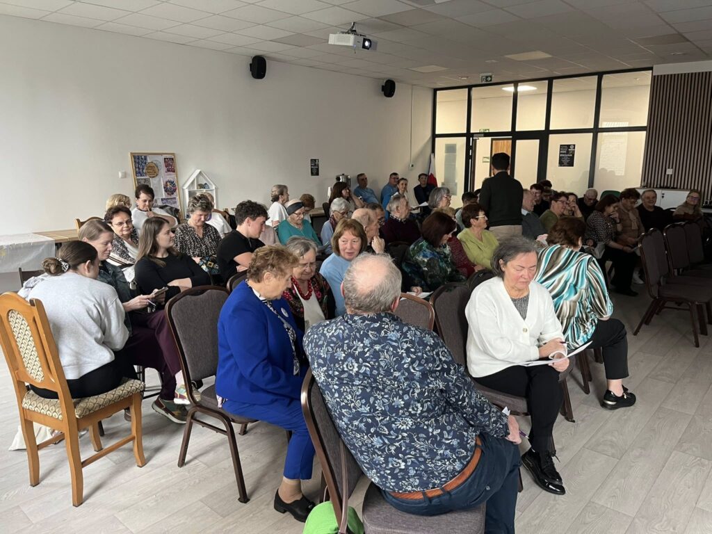 Grupa ludzi siedzi na krzesłach w jasnym pomieszczeniu z białymi ścianami. Uczestnicy rozmawiają między sobą, niektórzy trzymają notatki. W tle jest plakat i drewniane drzwi.