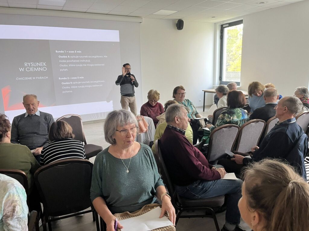 Grupa seniorów siedzi w sali szkoleniowej. Na ekranie pokazane są instrukcje ćwiczenia Rysunek w ciemno. Ujęcie przedstawia różnorodne interakcje i wymianę zdań.