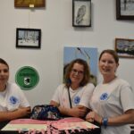 Trzy kobiety w białych koszulkach siedzą przy stole na tle obrazów. Na ścianie zielony znak Uniwersytet Dobrego Wieku. Na stole różowe wstążki i ulotki.