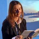 Osoba w garniturze siedzi i czyta książkę. W tle widać zdjęcia krajobrazów. Światło pada na twarz z lewej strony.