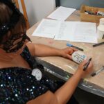 Na zdjęciu osoba w masce maluje napis na kamieniu. Ma na sobie cekinową suknię, przy stole leżą pędzle i kartki. Tło jest jasne, a na stole widać drewniane pudełko z przyborami.